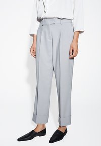 Person in hellgrauen maßgeschneiderten Hosen mit umgeschlagenen Säumen, weißer Bluse und schwarzen Slippern vor einem weißen Hintergrund.