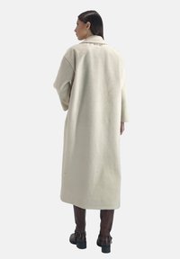 Ljusbeige lång rock av mjukt tyg med vida ärmar och sprund bak, kombinerad med bruna knähöga stövlar.