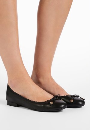 Czarne baleriny ze skóry z falistymi brzegami i małymi kokardkami, noszone na bose stopy na białym tle.