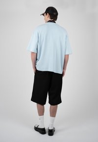 Homme portant une casquette noire, une chemise oversize bleu clair à manches courtes, un short noir, des chaussettes blanches et des chaussures noires, debout dos à nous sur fond blanc.