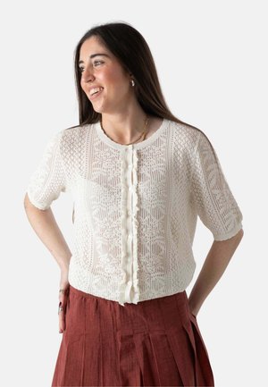 Mujer sonriendo, vestida con un top de botones de encaje blanco de manga corta y una falda plisada de color óxido, con las manos apoyadas en las caderas.