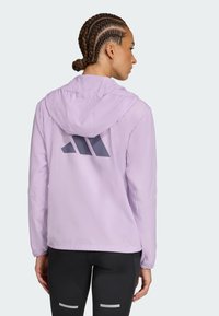 Veste à capuche lilas avec une texture lisse, des poignets élastiques et un logo gris bien visible au dos. Associée à des leggings noirs.