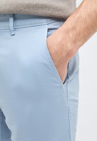 Ανδρικό χέρι που είναι τοποθετημένο στην εμπρόσθια τσέπη ανοιχτόχρωμων μπλε παντελονιών, φορώντας μπεζ πλεκτό πουλόβερ.