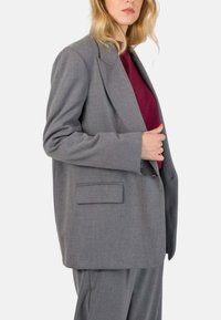 Blazer grigio oversize realizzato in tessuto testurizzato, con revers a punta, tasca singola e fit ampio, indossato sopra un top bordeaux.