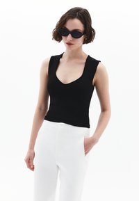 Top negro sin mangas con escote en forma de barco, combinado con pantalones blancos de talle alto que tienen bolsillos laterales y costuras verticales.