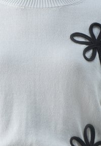 Hellgrauer gestrickter Pullover mit schwarzen Stoffblumen-Applikationen auf der linken Seite. Die Textur wirkt weich und hat einen gerippten Halsausschnitt.