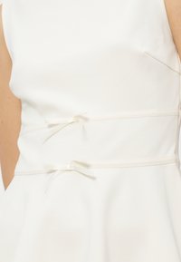 Közelkép egy fehér ujjatlan ruháról, amelyen két vékony derékszíj kis masnira kötve oldalt.