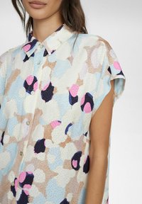 Camicetta a maniche corte in tessuto leggero con un motivo astratto multicolore in diverse tonalità di blu, rosa e beige. Chiusura frontale con bottoni, dettaglio del colletto.