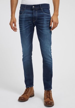 Schmal geschnittene dunkelblaue Jeans, getragen mit braunen Leder-Schnürstiefeln bei einer stehenden Person vor einfarbigem Hintergrund.
