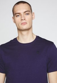 Camiseta corta de manga en color púrpura, hecha de una tela suave, con un escote redondo y un discreto logotipo negro en el pecho. Textura suave.