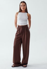 White ribbed tank top with side ties paired with brown wide-leg trousers. Outfit features a smooth texture and a relaxed fit. Black flat sandals.