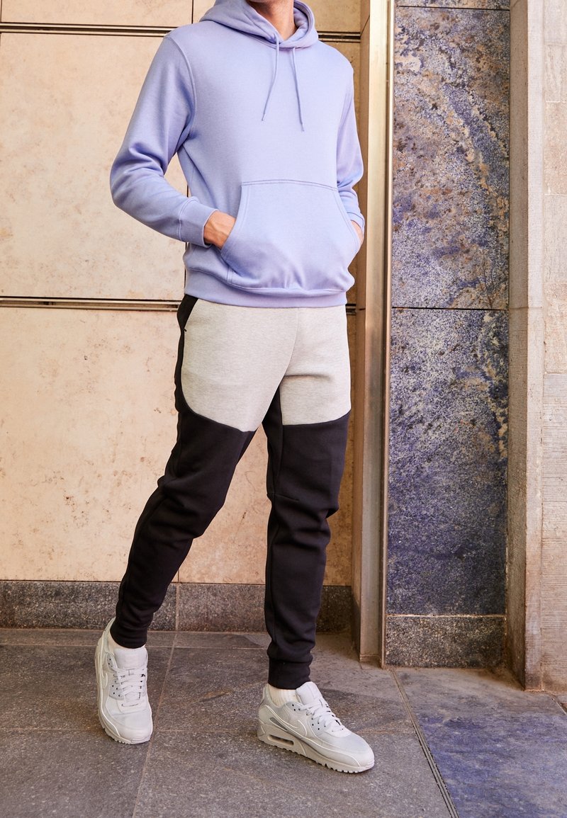 Ljuslila huva-tröja med en framficka, parat med grå och svart färgblockerade joggers och ljusgråa träningssneakers.