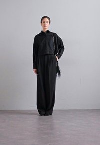Bluză neagră scurtă cu model texturat, asortată cu pantaloni negri largi. Modelul ține o geantă mică cu franjuri. Look simplu, monocrom.