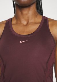 Canotta sportiva color marrone scuro con design aderente, caratterizzata da una texture liscia e un piccolo logo Nike rosa sul petto.