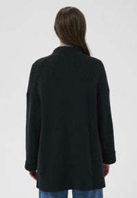 Person mit langen braunen Haaren, der eine schwarze, gerippte Strickjacke und eine blaue Jeans trägt, steht mit dem Rücken zu einer schlichten Hintergrund wand.