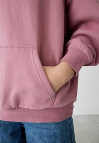Felpa rosa con tasca frontale e polsini a coste, realizzata in un morbido tessuto testurizzato. Abbinata a jeans in denim blu, con una leggera sfumatura.