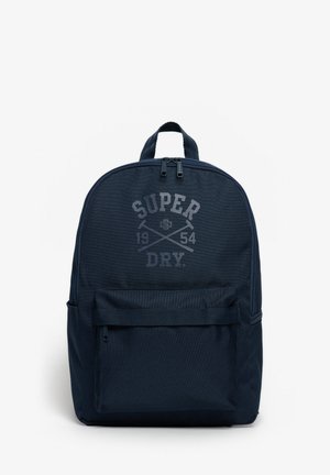 Sac à dos bleu foncé avec poche zippée avant et logo texte "Super Dry 1954" sur le devant sur fond blanc.
