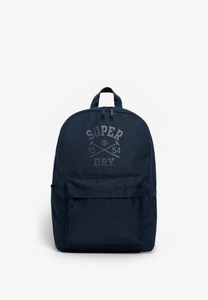 Ciemnoniebieski plecak z przednią kieszenią na zamek i napisem logo "Super Dry 1954" z przodu na białym tle.