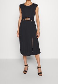 Černé šaty po kolena s přiléhavým korzetem, krátkými rukávy a páskem v pase s dvěma hnědými popruhy. Obsahují boční rozparek pro snadnější pohyb.