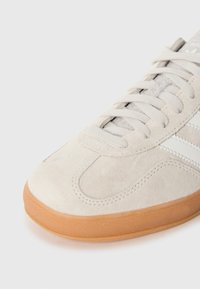 Detail béžovej semišovej tenisky s bielymi pruhmi a podrážkou z gumy na bielom pozadí.