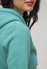 Sudadera con capucha turquesa de tejido texturizado y moteado; presenta un diseño relajado y detalles de costura visibles en la capucha y el hombro.