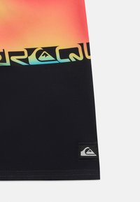 Tissu noir avec une bande dégradée orange à jaune en haut, présentant un texte "Quiksilver" stylisé en bleu et jaune ainsi qu'un patch logo à l'ourlet.