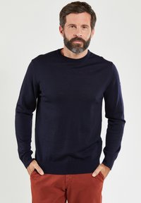 Navyblauw gebreid trui met ronde hals, lange mouwen, ribgebreide manchetten en onderrand, effen kleur, zachte textuur, gedragen over roestkleurige broeken.