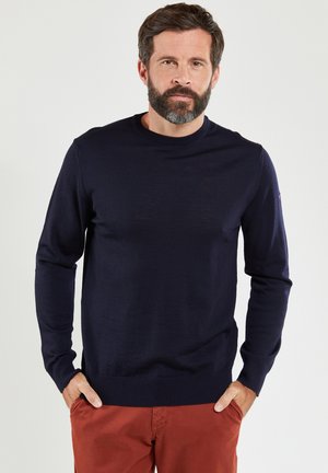 Marineblauer Strickpullover mit Rundhalsausschnitt, langen Ärmeln, gerippten Bündchen und Saum, einfarbig, weiche Textur, getragen über rostfarbenen Hosen.