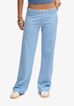 Person iført lyseblå bukser med vide ben med et Superdry-mærke ved taljen og hvide sneakers, trækker let i taljen med hånden.