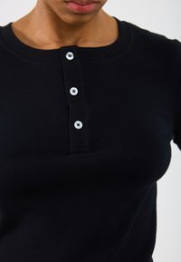 Črna rebrasta majica z okroglo izrezom, trojnim gumbom z belimi gumbi in oprijetim dizajnom, ki poudarja obline.