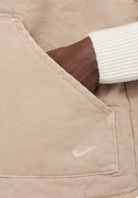 Bežs auduma žaketes kabata ar vieglu tekstūru. Redzama roka kabatā, ar baltu ribotu piedurkni. Izšūta Nike logo.