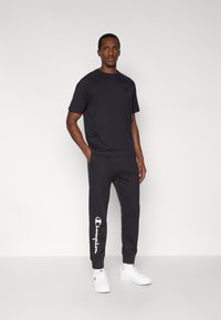 Μαύρο κοντομάνικο μπλουζάκι συνδυασμένο με σκούρα παντελόνια φόρμας που φέρουν λευκό λογότυπο «Champion». Τα παντελόνια φόρμας έχουν άνετη εφαρμογή και ελαστικά μανσέτα.