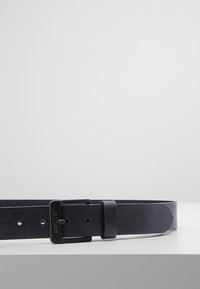 Ceinture en cuir noir avec une finition mate, boucle rectangulaire et plusieurs trous pour le réglage de la taille. Présentée sur un fond clair.