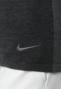 Grå stickad polo med en texturerad yta, som har en liten silverfärgad Nike-logga på vänster sida och en slät vit tygdel nederst.