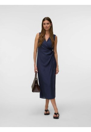 Femme en robe portefeuille bleu marine sans manches, tenant un sac à main marron, portant des sandales noires, debout sur un fond clair uni.