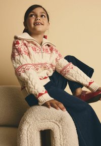 Krämfärgad kabelstickad tröja med röda dekorativa mönster, med dragkedja vid kragen, kombinerad med marinblå jeans och röda sandaler, sittande på en strukturerad soffa.