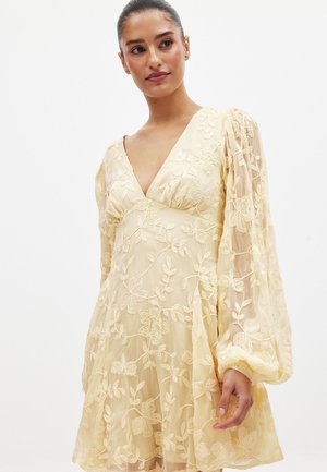 Geltonas gėlėmis siuvinėtas suknelė su gilia V iškirpte, permatomomis ilgomis rankovėmis ir susiraukšlėjusia juosena, turinti išraiškingus lapų ir gėlių raštus.