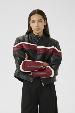 Woman with long brown hair wearing black leather jacket with red and white stripes, standing with arms crossed against neutral background.