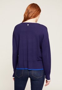 Lila stickad tröja med rund halsringning, långa ärmar och en kontrasterande nederkant i blått och vitt. Har en liten logotyp på baksidan.