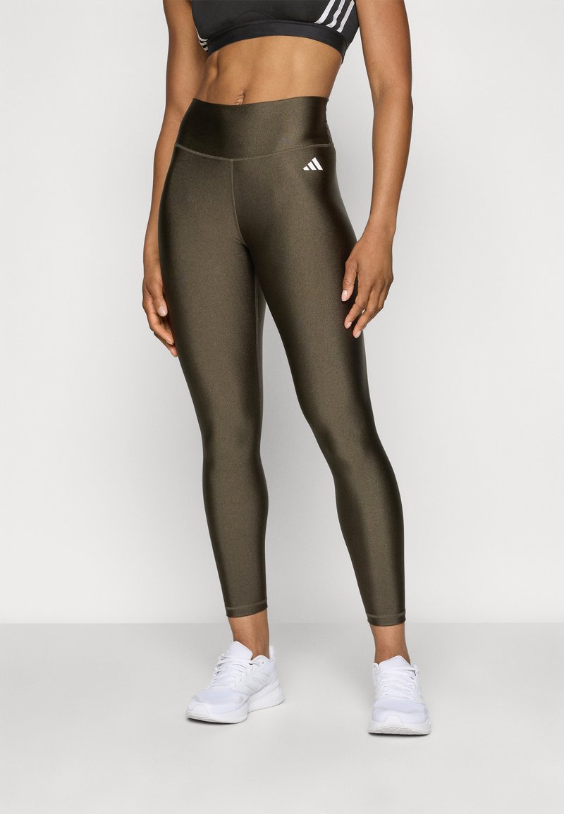 Frau in glänzenden olivgrünen High-Waist-Leggings mit weißem Logo, schwarzem Sport-BH und weißen Sportschuhen, die vor einem schlichten Hintergrund steht.