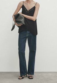 Femme portant un caraco noir orné de dentelle, un jean bleu foncé, des sandales à talons noires, tenant une grande pochette noire.