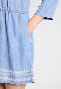 Personne portant une robe bleu clair avec un motif brodé blanc au ourlet, la main droite placée dans la poche latérale.