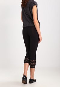 Svart capri leggings med spetsdetalj vid nederkanten, kombinerat med en grå t-shirt med kort ärm och svarta skor. Designen är figurnära och avslappnad.