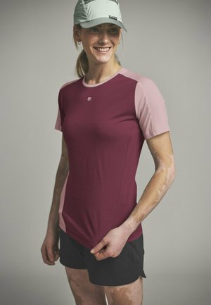 Femme souriante portant un t-shirt de sport bicolore bordeaux et rose, un short noir et une casquette gris clair, debout sur un fond uni.