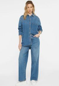 Denim shirt jacket with button front, large pocket, and long sleeves; paired with wide-leg denim pants and white sneakers.