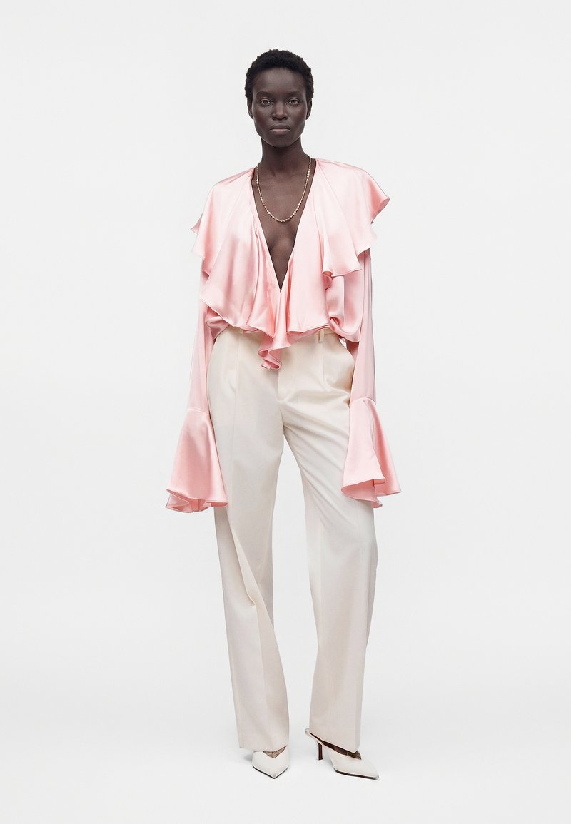 Femme portant un chemisier en satin rose clair à volants avec des manches larges, un pantalon taille haute crème, des talons blancs et un collier chaîne en or.