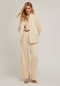 Costum din material satin de culoare crem, cu un sacou ajustat și pantaloni evazați. Include un închidere cu un singur nasture și buzunare laterale.
