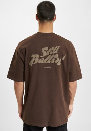 Brun bomulls t-skjorte med stort, preget "Still Ballin'" grafikk i lys farge på baksiden, kortermet og en avslappet passform.