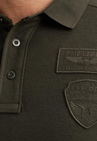 Maslinasto zeleni polo majica od teksturirane, rebraste tkanine; sadrži dvije crne dugmadi i dvije izvezene oznake na prsima.