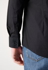 Svart langermet skjorte med knapper på ermene, laget av glatt stoff, sammen med lyseblå denimjeans med baklomme.